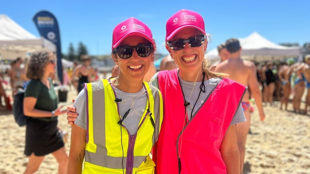 Two volunteer coordinators in hi-vis vests and event caps, radios clipped on, at the registration area on the beach before an ocean swim